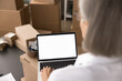 © fizkes - Woman working at desk covered with various packages, white mock up laptop screen view over female shoulder. Logistics, inventory management, e-commerce operations, packages for dispatch preparation