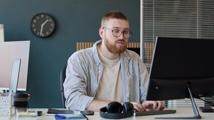 Medium portrait of young bearded Caucasian man wearing smart casual clothes looking at camera while working on computer