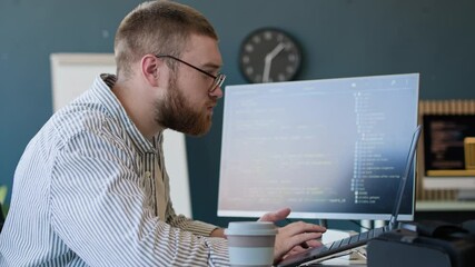 Side view of young Caucasian programmer typing code on laptop and checking data on desktop computer and in notebook