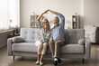 © fizkes - Happy senior homeowners couple sit on sofa in modern living room, joining their hands showing roof over their heads, looking at camera, celebrate relocation day to new house, long-term rent, bank loan