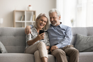 Happy aged wife and husband sit on sofa look at mobile phone screen, laughing, watching funny videos in social media, spend carefree pastime on internet using gadget. Olde gen and modern tech usage