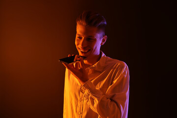 Poster - Young man recording voice message via smartphone on color background with neon lights