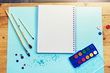 Notebook, paint and above with brushes for creative workspace, art or design on desk. Top view of empty space with paper, poster and color palette for activity or creativity on wooden table or mockup