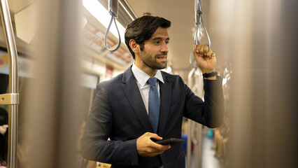 Wall Mural - Skilled business man standing in train or subway while holding phone. Professional project manager looking around while enjoy in train ride while playing mobile phone at train station. Exultant.