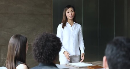 Poster - Serious young Asian business coach giving lecture, seminar, management class to diverse team. Project manager woman discussing creative ideas with colleagues, holding brainstorming meeting