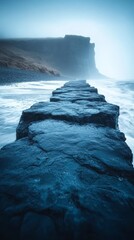 Sticker - Rocky path extending into misty ocean water, leading towards a distant, towering cliff under a moody sky