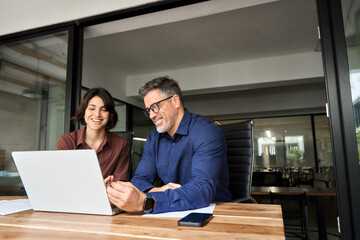 Wall Mural - Male executive manager explaining corporate online project plan to female partner sitting at office desk. Two happy busy professionals business team people working together using laptop computer.