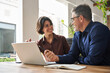 © insta_photos - Two busy business team people using laptop computer working together sitting at office meeting. Happy young woman and mature man entrepreneurs or executives smiling at work talking in modern workspace