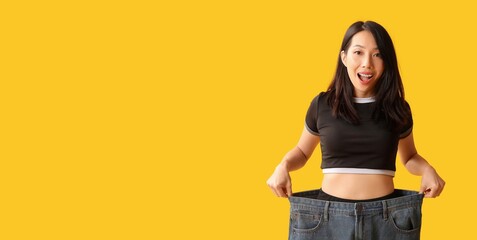 Poster - Happy young Asian woman in loose jeans on yellow background with space for text. Weight loss concept