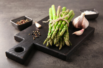 Board with bunch of fresh green asparagus stems, garlic and peppercorns on grey textured table, closeup