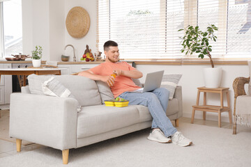 Canvas Print - Young man with laptop and lunch box opening bottle of juice at home