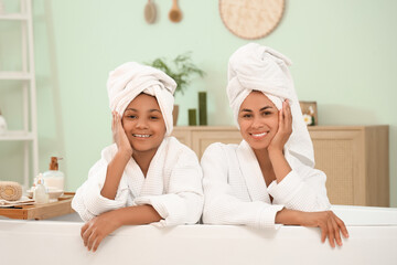 Wall Mural - Happy young African-American mother and her daughter in bathrobes sitting inside bathtub at home