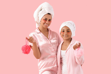 Poster - Happy young African-American mother and her daughter in pajamas with under-eye patches holding bath sponges on pink background