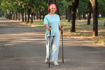 Wall Mural - Mature woman in headphones training with walking poles in park