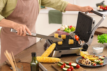 Young man cooking fresh corn cobs on modern electric grill in kitchen, closeup