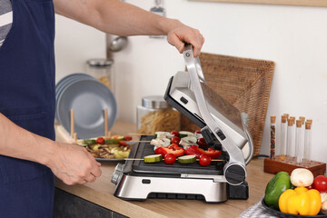 Young man cooking tasty fresh vegetables skewers on modern electric grill in kitchen, closeup