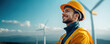 © khonkangrua - A smiling engineer wearing a yellow helmet and safety gear, standing outdoors near wind turbines, symbolizing renewable energy and sustainability.