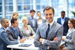 © Tanicsean - A professional man in a gray suit, smiling warmly, stands with his arms crossed while colleagues engage in a group discussion in a contemporary office