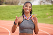 © Nina2024/peopleimages.com - Woman, celebration and thumbs up with medal as athlete for win, competition and sports achievement outdoor. African person, portrait and success with professional running, award or podium performance