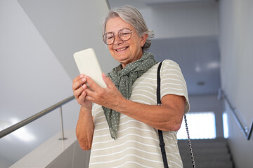 Wall Mural - Portrait of smiling mature attractive caucasian gray haired woman walking down  stairs inside a building using mobile phone