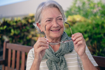 Wall Mural - Portrait of happy attractive senior woman with short gray hair sitting outdoor in public park holding her eyeglasses. Smiling elderly caucasian lady enjoying free time and good day