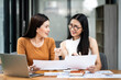 © NAMPIX - Two asian business women sit at desk discuss project details, share opinion, working on collaborative task