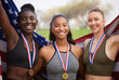 © Nina2024/peopleimages.com - Women, portrait and sport medal with USA flag, achievement and team running award outdoor. Marathon, trophy and stadium win with runner group and smile from competition relay success with celebration