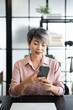 © Fahng - Middle-aged Asian Businesswoman stting in home office with a mobile phone in hand.Small business entrepreneur looking at mobile phone while communicating with colleagues in office.