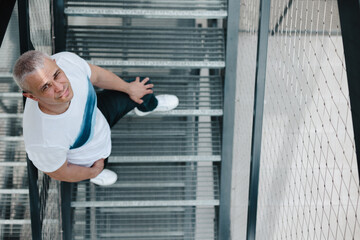 A man is standing on a set of stairs with a smile on his face. He is wearing a white shirt and blue jeans