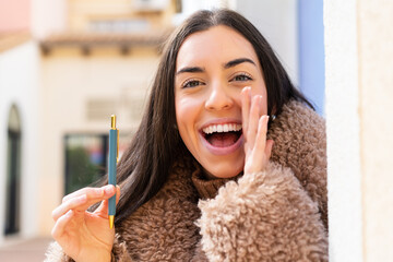 Wall Mural - Young woman holding a pen at outdoors shouting with mouth wide open