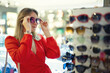 © M-Production - A woman in a red dress tries on glasses in a store.