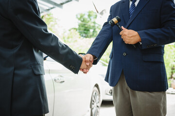 Two Asian individuals discuss a car insurance claim with an agent in front of a damaged vehicle. They review documents, discuss the policy, and address the crash and repair costs.