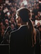 © vefimov - A woman stands behind a microphone in front of a crowd. She is wearing a suit and has her hair in a ponytail. Concept of professionalism and authority