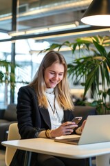 Wall Mural - Young woman in business attire smiling, using a tablet and laptop at a desk in an office environment.