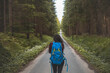 © Fauren - Back view of a woman with a blue backpack walking barefoot down a peaceful forest road, surrounded by lush greenery and tall trees, symbolizing adventure and tranquility in nature