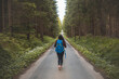 © Fauren - Back view of a woman with a blue backpack walking barefoot down a peaceful forest road, surrounded by lush greenery and tall trees, symbolizing adventure and tranquility in nature