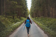 © Fauren - Back view of a woman with a blue backpack walking barefoot down a peaceful forest road, surrounded by lush greenery and tall trees, symbolizing adventure and tranquility in nature