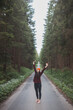 © Fauren - Smiling woman in a red checkered shirt and bright orange beanie stands in the middle of a forest road, stretching her arms behind her head, enjoying the tranquility of the surroundings
