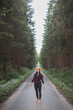 © Fauren - Smiling woman in a red checkered shirt and bright orange beanie stands in the middle of a forest road, stretching her arms behind her head, enjoying the tranquility of the surroundings