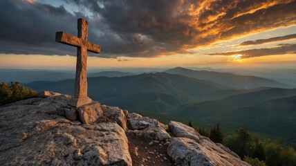 A wooden cross stands atop a rocky peak against a vibrant sunset in the mountains, illuminated by warm orange and purple hues during twilight