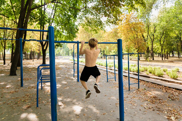 View from the back as a teenager in the park pulls himself up on the bar.  Athletic teenage boy in the park outdoor workout session