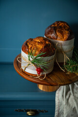 Two homemade Christmas panettone, made as holiday gift with golden deer and pine and red berries decorations on wooden cake stand on blue wooden cupboard .