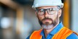 © ABULKALAM - An image of a guy engineer or construction worker standing with his arms crossed, wearing an orange safety vest, protective glasses, and a white hard hat. The man appears serious.