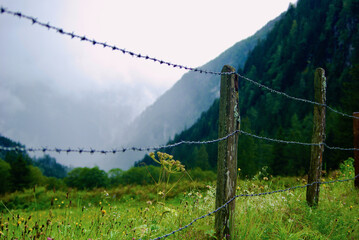 Wall Mural - view of an mountain landscape with a meadow fenced with barbed wire on a rainy day with gray clouds 