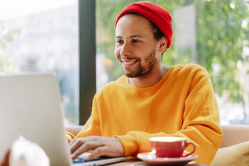 Smiling young man wearing stylish red hat working on laptop in cafe