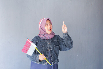 A woman in a purple hijab feels happy while holding a red and white flag. The red and white flag is the national flag of Indonesia.