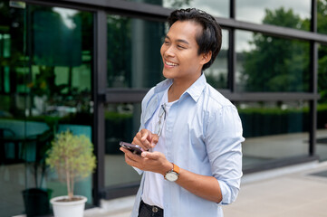 Wall Mural - A positive Asian man is walking along a building in the city with a smartphone in his hand.