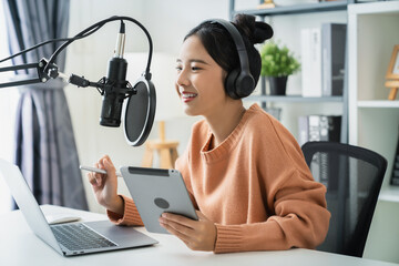 Young Asian woman holding digital tablet and use microphones wear headphones with laptop record podcast interview for radio. Content creator concept.