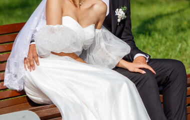 The bride in a wedding dress and the groom are sitting on a bench in the park. Detailed photo