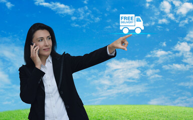 Wall Mural - Businesswoman talking on her cell phone and pointing finger to free delivery truck icon over green grass field with blue sky, Business transportation online service concept